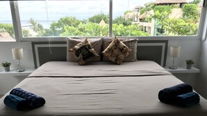 Una cama bien hecha con vistas al océano a través de amplias ventanas, dos lámparas de noche y una pequeña planta en maceta sobre la mesita.