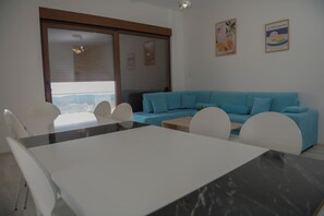A modern living room with a blue sofa, white chairs, a dining table, and a large window with blinds.