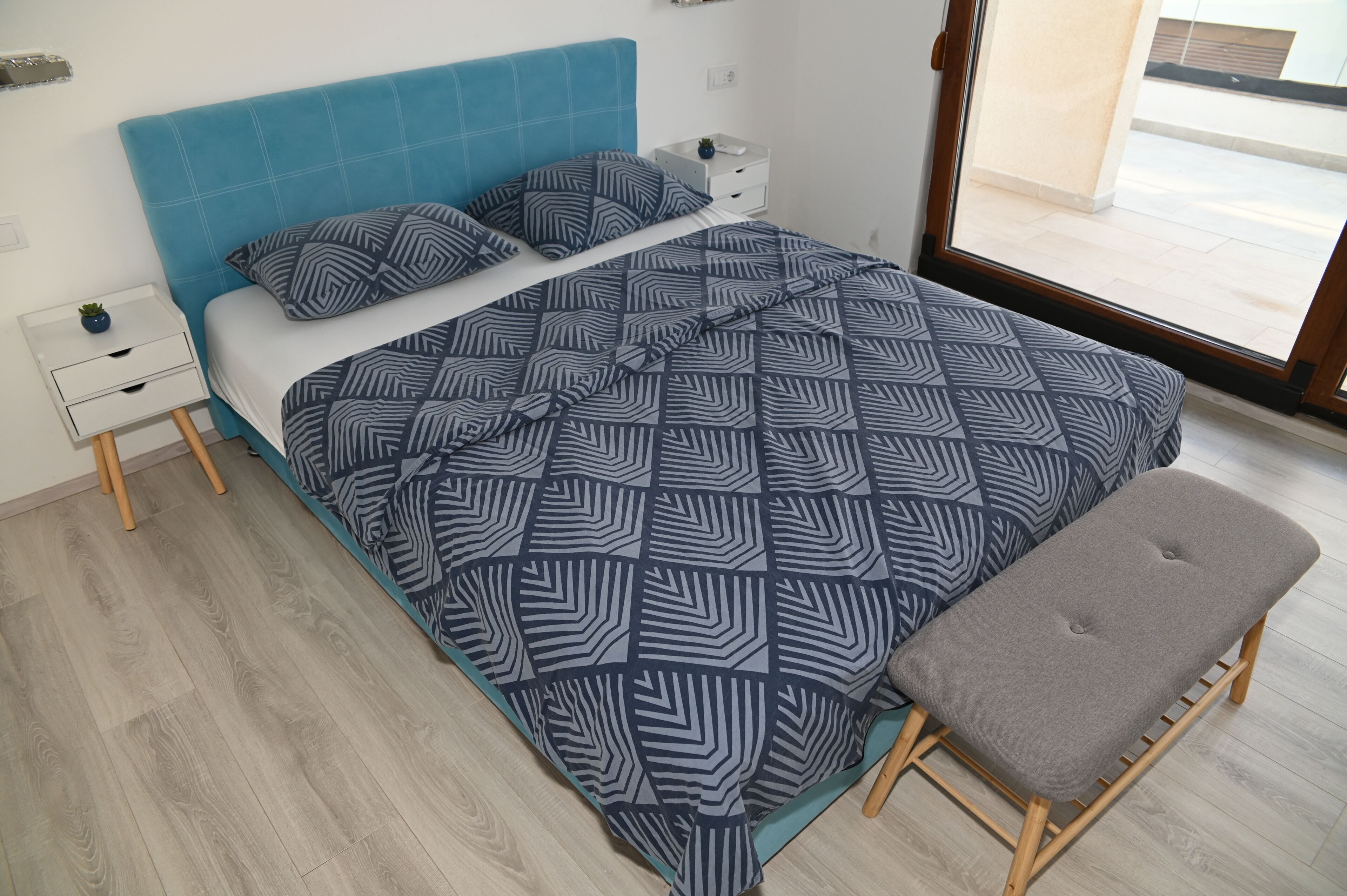 Ein Bett mit geometrisch gemustertem Überwurf, ein passendes Kopfkissen, ein Nachttisch mit Pflanze und eine Bank.
