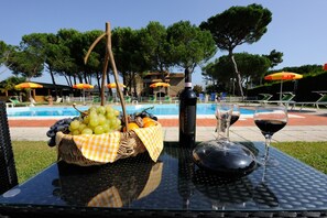 Une table avec une bouteille de vin, des verres et un panier de fruits, placée au bord d’une piscine.