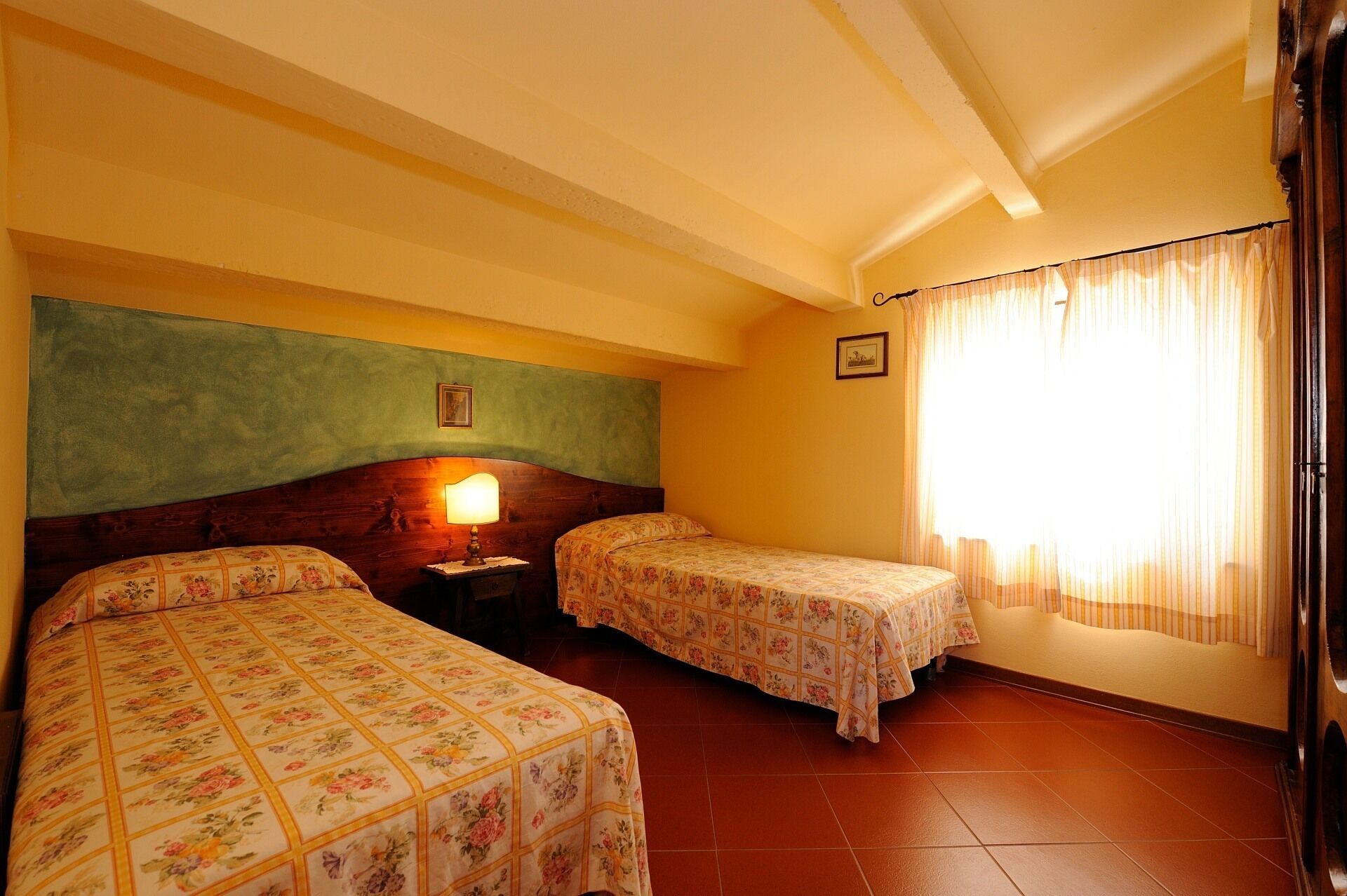 A room with two beds, a nightstand, a lamp, a window with curtains, and a picture on the wall.