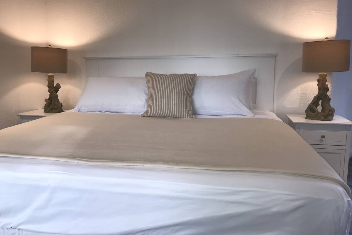 A neatly made bed with white linens and two pillows, flanked by two bedside tables with lamps.