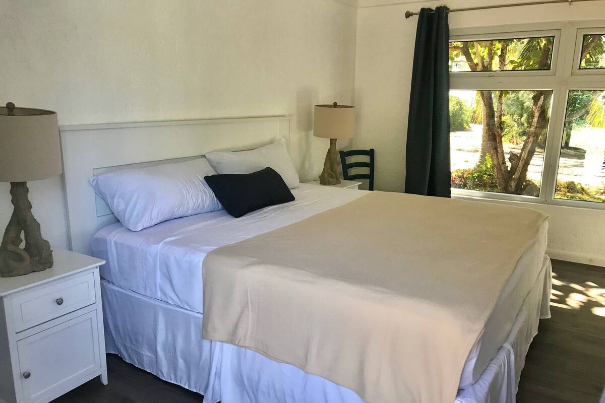 A neatly made bed with a beige blanket, white bedding, and a black pillow. There is a nightstand with a lamp and a drawer. A chair is placed near a window with a view of trees.