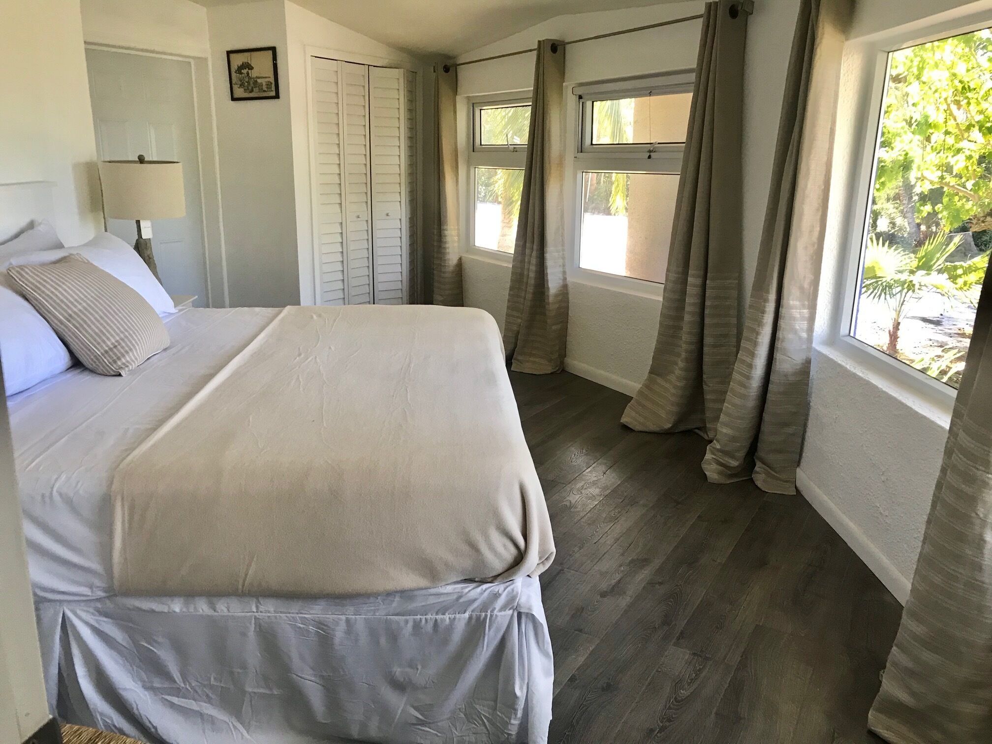 A neatly made bed with a white comforter and pillows, a bedside lamp, a window with curtains, and a view of greenery outside.