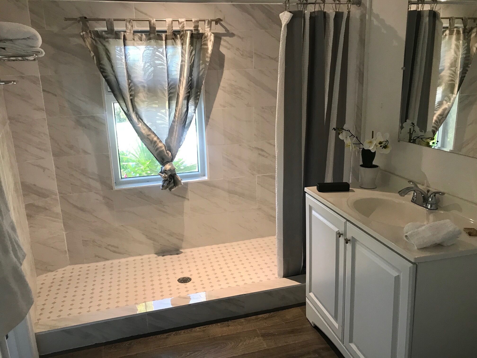 A modern bathroom with a walk-in shower, a window with sheer curtains, a sink with a mirror, and a towel rack.