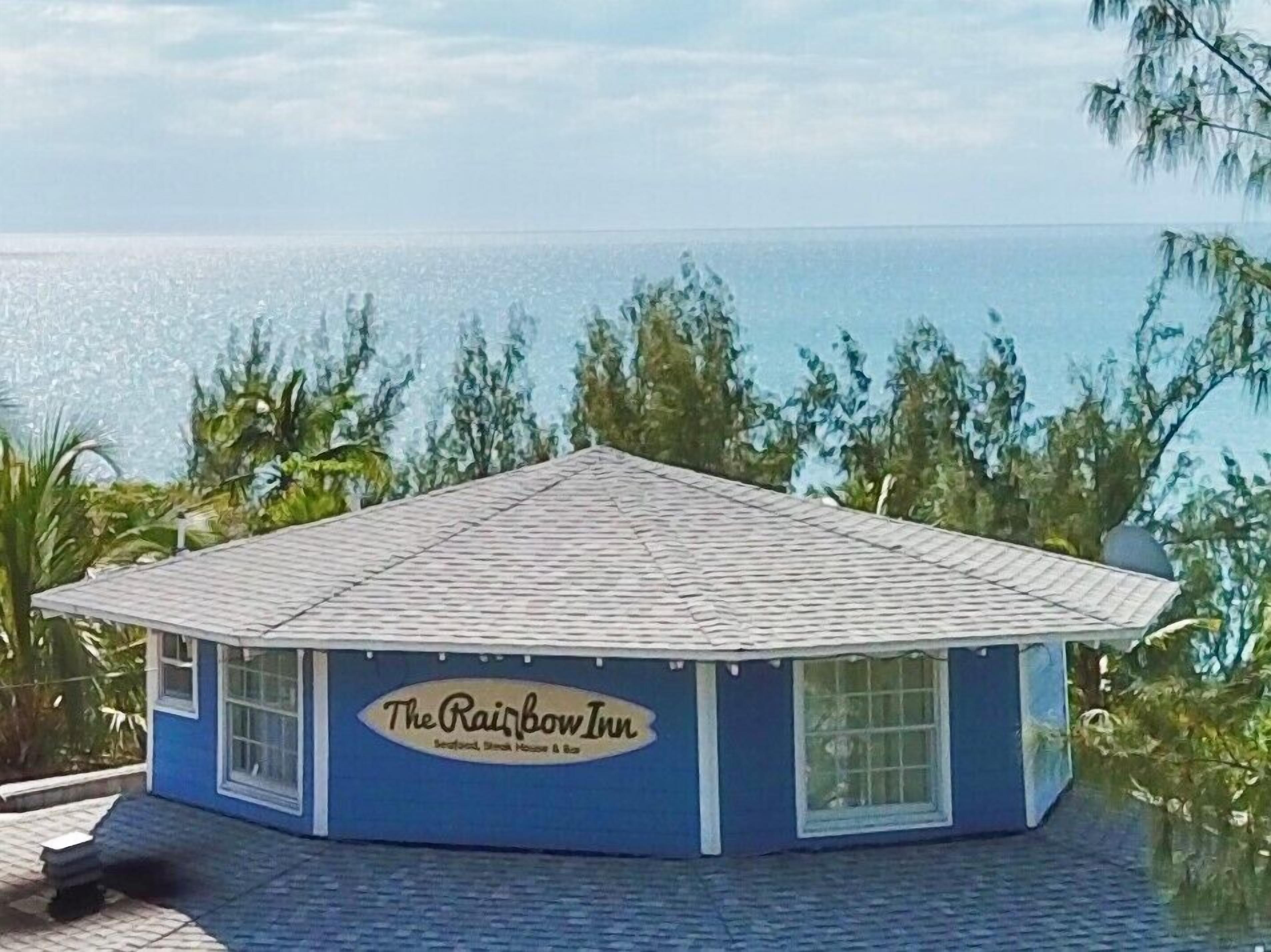 Ein kleines, eingeschossiges Gebäude mit blauer Fassade und einem Schild, auf dem "The Rainbow Inn" steht. Es hat ein geneigtes Dach und wird von Grünpflanzen umgeben, mit Blick auf den Ozean.