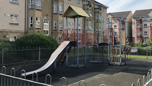 A playground with a slide and climbing structures, surrounded by a metal fence, with residential buildings in the background. - McDonald Road Apartment (Edinburgh)