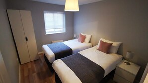 A small, well-lit bedroom with two beds, a window, a wardrobe, and a bedside table with a lamp. - McDonald Road Apartment (Edinburgh)