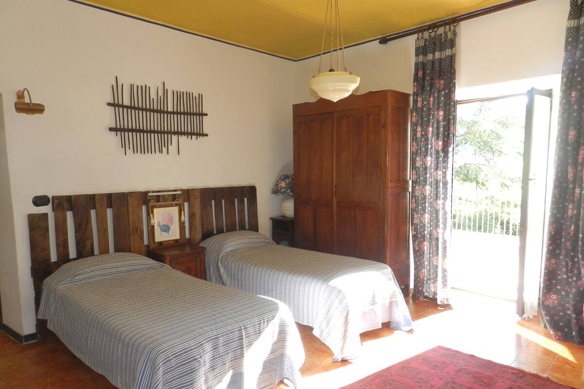 A room with two beds, a wooden wardrobe, a hanging light fixture, and a view of the outdoors through a window with curtains.