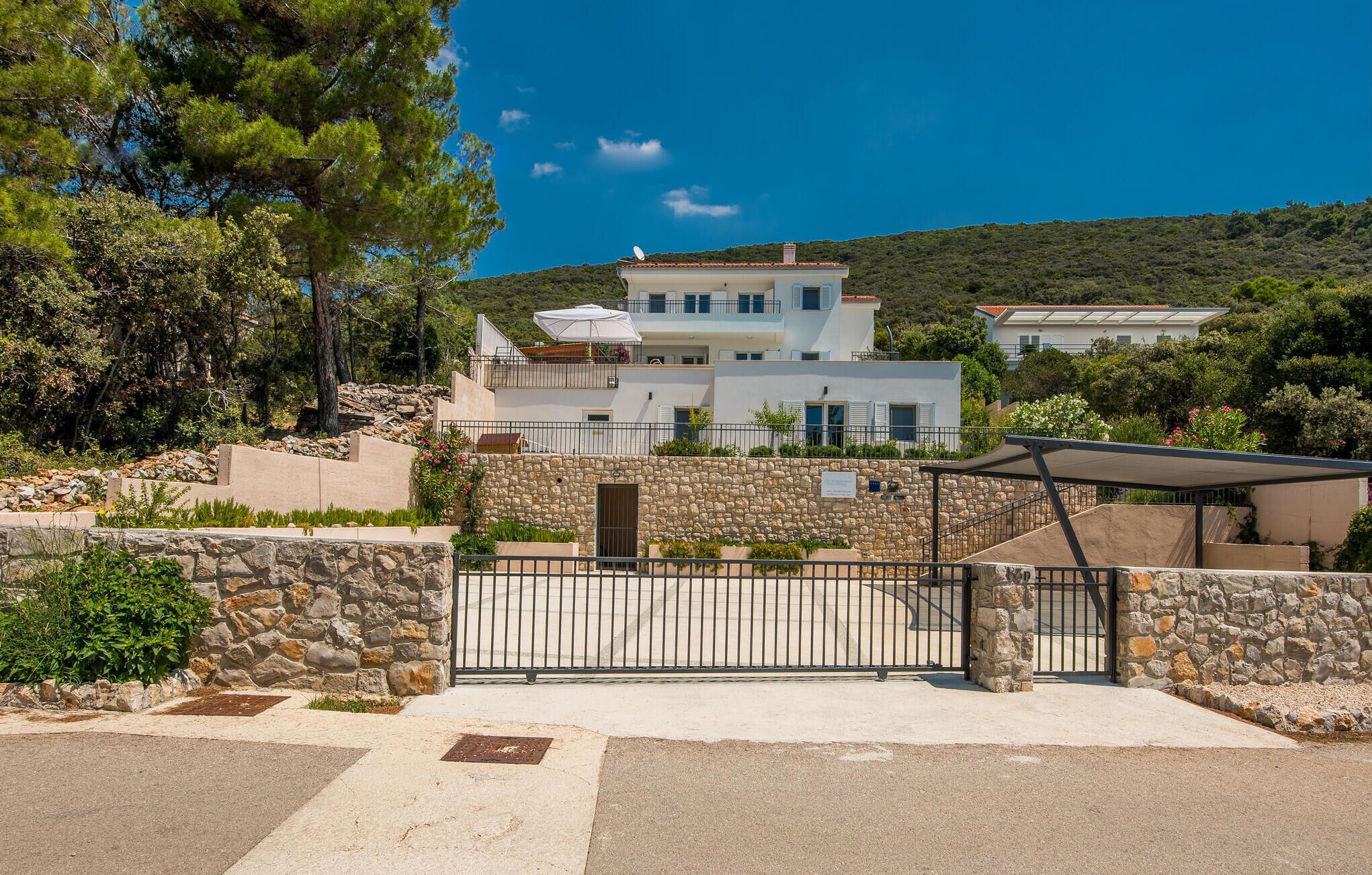 Une maison moderne à deux étages, dotée d’un mur en pierre et d’un portail en métal, entourée de verdure et surplombant un ciel bleu azur.