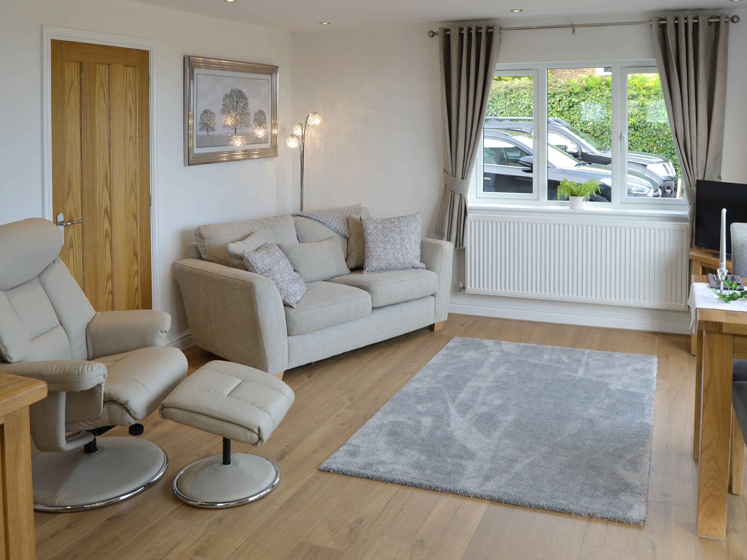 Ein Wohnzimmer mit einer Couch, einem Sessel und einem Hocker, einem grauen Teppich, einer Heizung, einem Fenster mit Vorhängen und einem gerahmten Bild an der Wand.