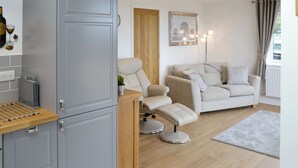 Ein modernes Wohnzimmer mit einer Couch, einem Liegestuhl, einem Holztisch und einer Küchenzeile mit Schränken und einem Wäscheständer.