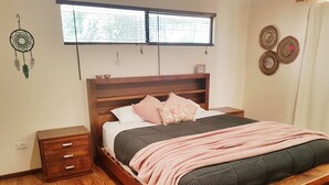 Una cama de madera con colcha gris y negra, una manta rosa y una mesita de noche de madera con cajones.