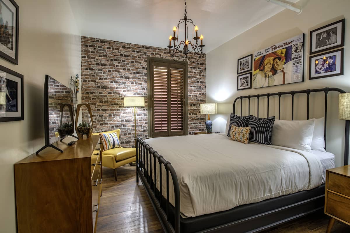 A bedroom with a brick wall, a bed with white bedding, a wooden dresser, a yellow armchair, and a chandelier.