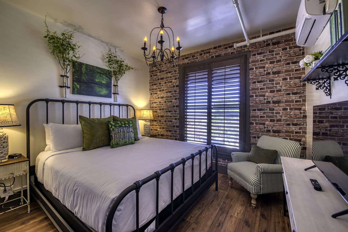 A bedroom with a brick wall, a bed with white bedding, a chandelier, a window with blinds, a chair, and a desk with a remote control.