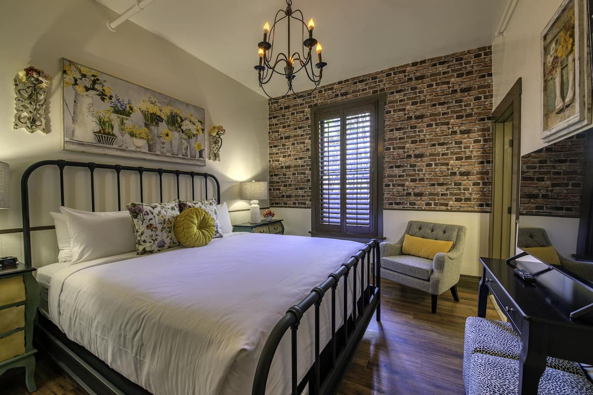 A bedroom with a brick wall, a bed with white bedding, a chandelier, and a chair with a yellow cushion.