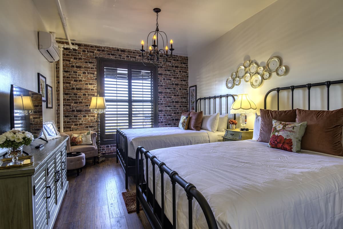 A bedroom with two beds, a brick wall, a chandelier, and a bedside table with a lamp.