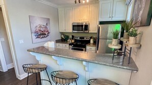 A modern kitchen with white cabinets, a stainless steel refrigerator, and a built-in microwave. There are three bar stools with metal frames and wooden seats.