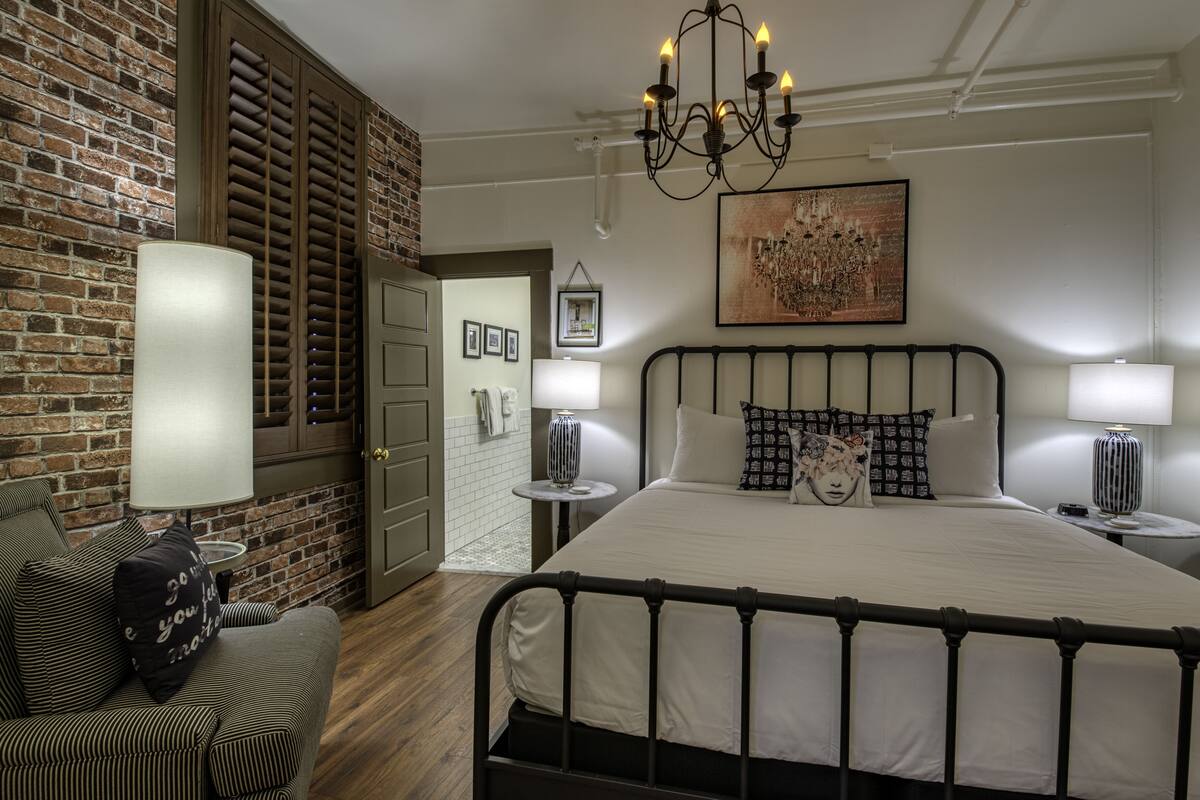 A bedroom with a large bed, a chandelier, a brick wall, and a door leading to another room.