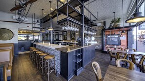 A modern restaurant interior with a central bar area, high ceiling with exposed beams, and a mix of bar stools and dining tables.