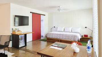 A hotel room with a bed, a desk with a refrigerator, a TV mounted on the wall, and a window with blinds.