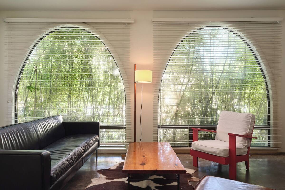 A modern living room with a leather sofa, a wooden coffee table, a red chair, and a cowhide rug. Large arched windows provide views of greenery outside.