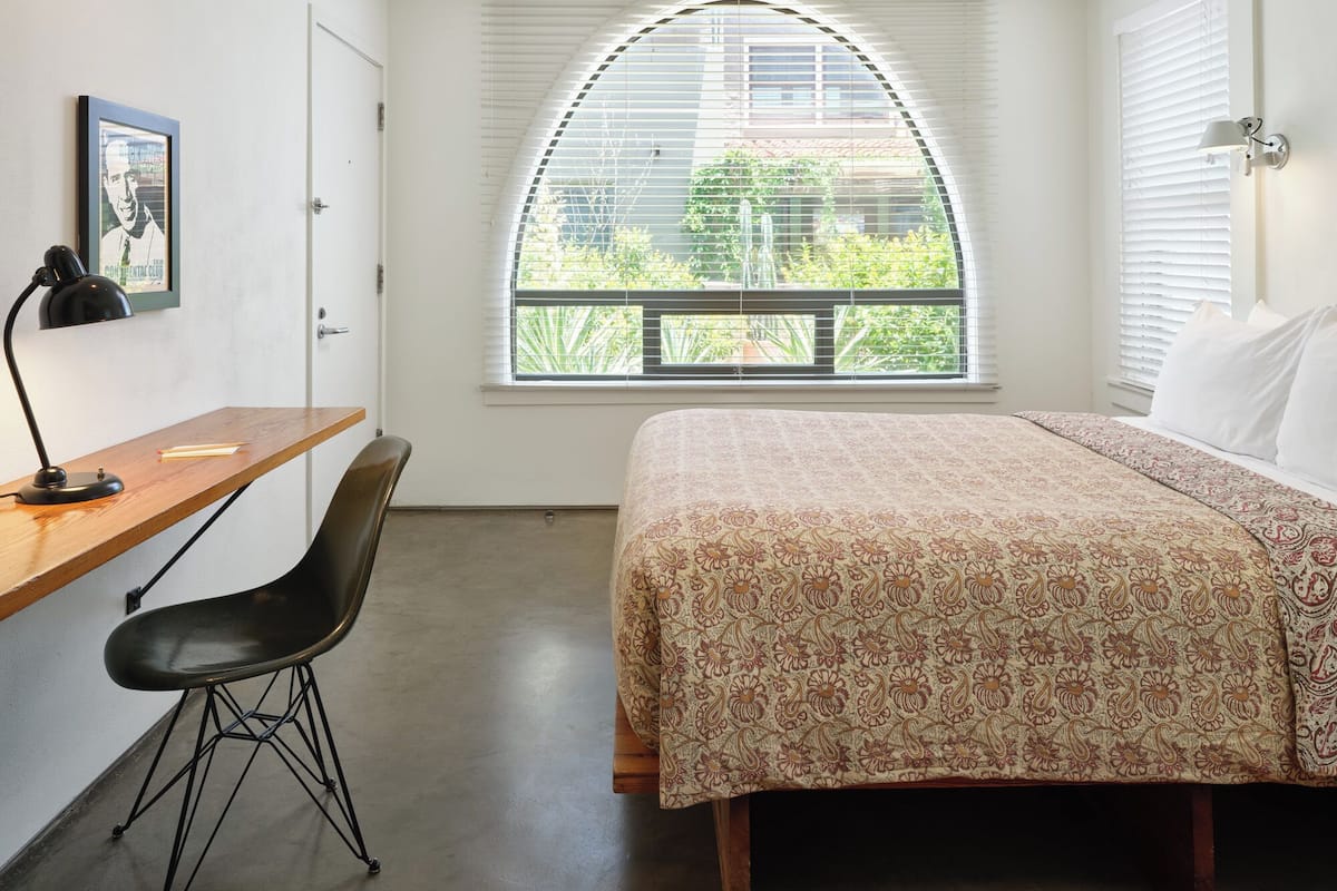 A bedroom with a bed, a desk with a chair, a window with blinds, and a framed picture on the wall.