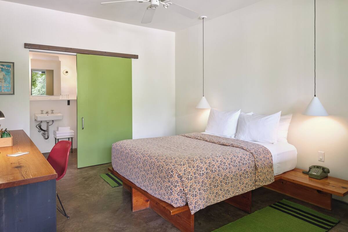 A bedroom with a bed, bedside tables, a desk, a chair, a green door, and a bathroom with a sink and mirror.