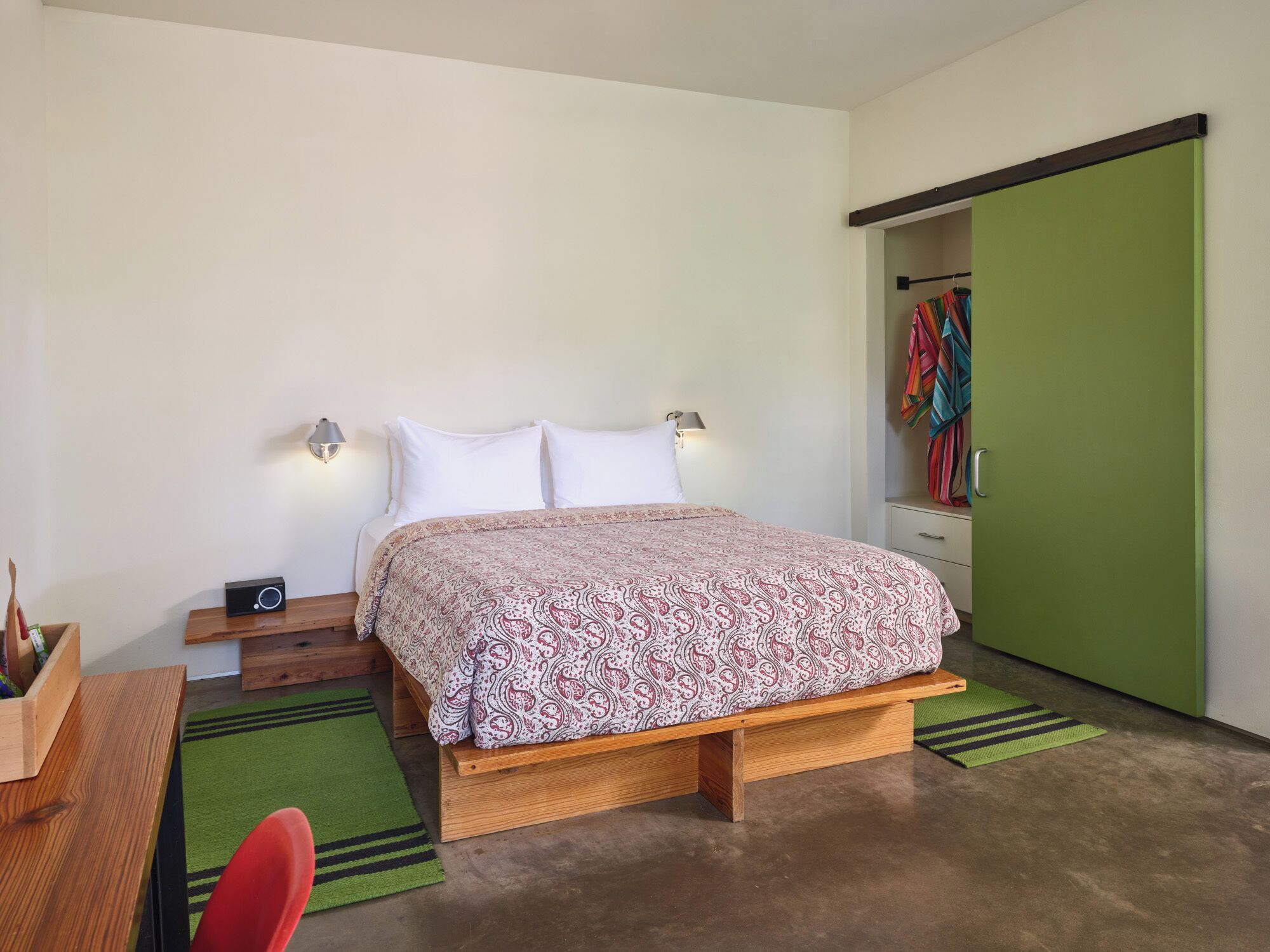 Un dormitorio con cama, lámparas de noche, una mesita de noche de madera, un escritorio, una silla, una puerta verde y un armario con ropa colorida.