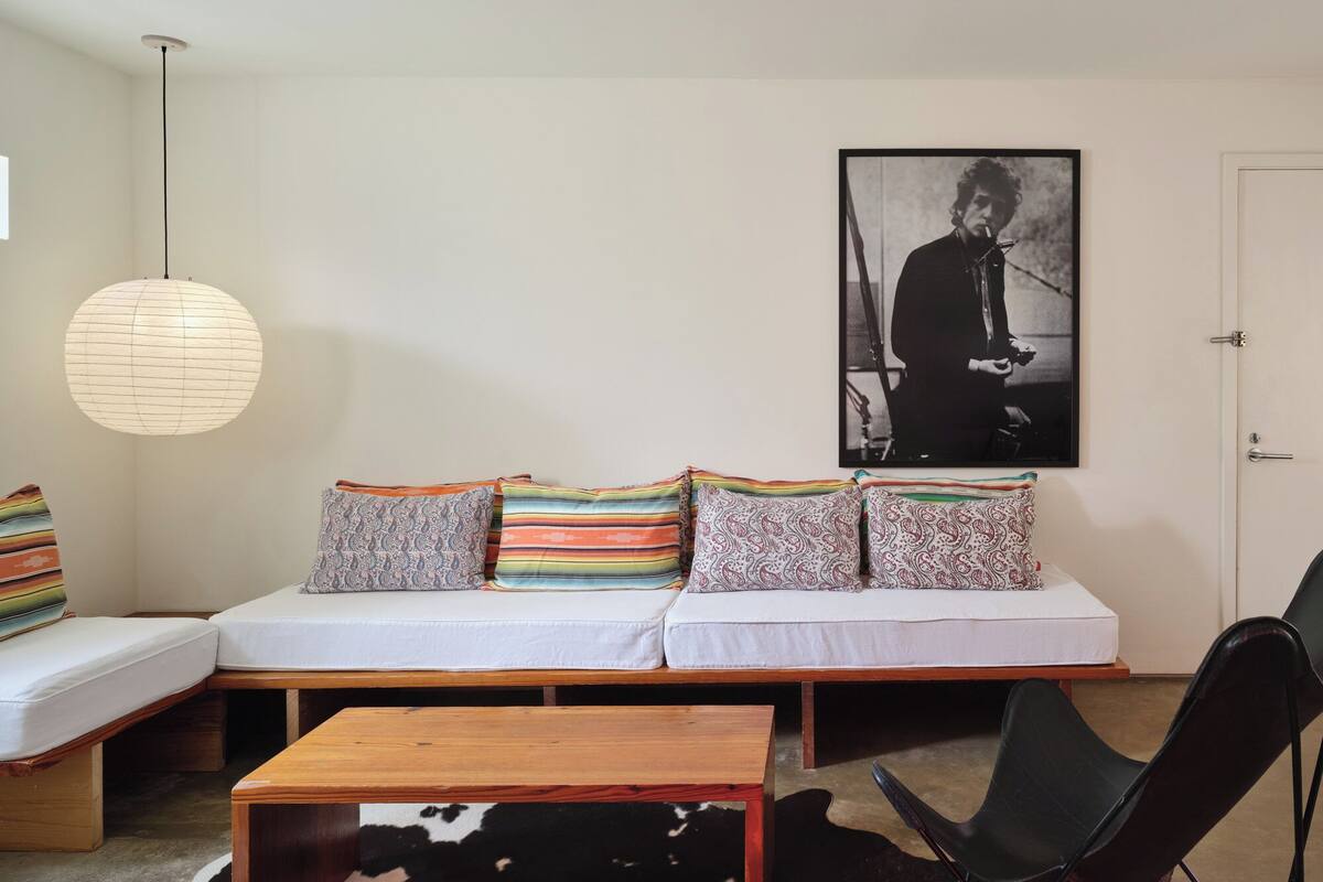 A modern living room with a sofa, patterned cushions, a wooden coffee table, a black chair, a hanging light fixture, and a framed picture on the wall.