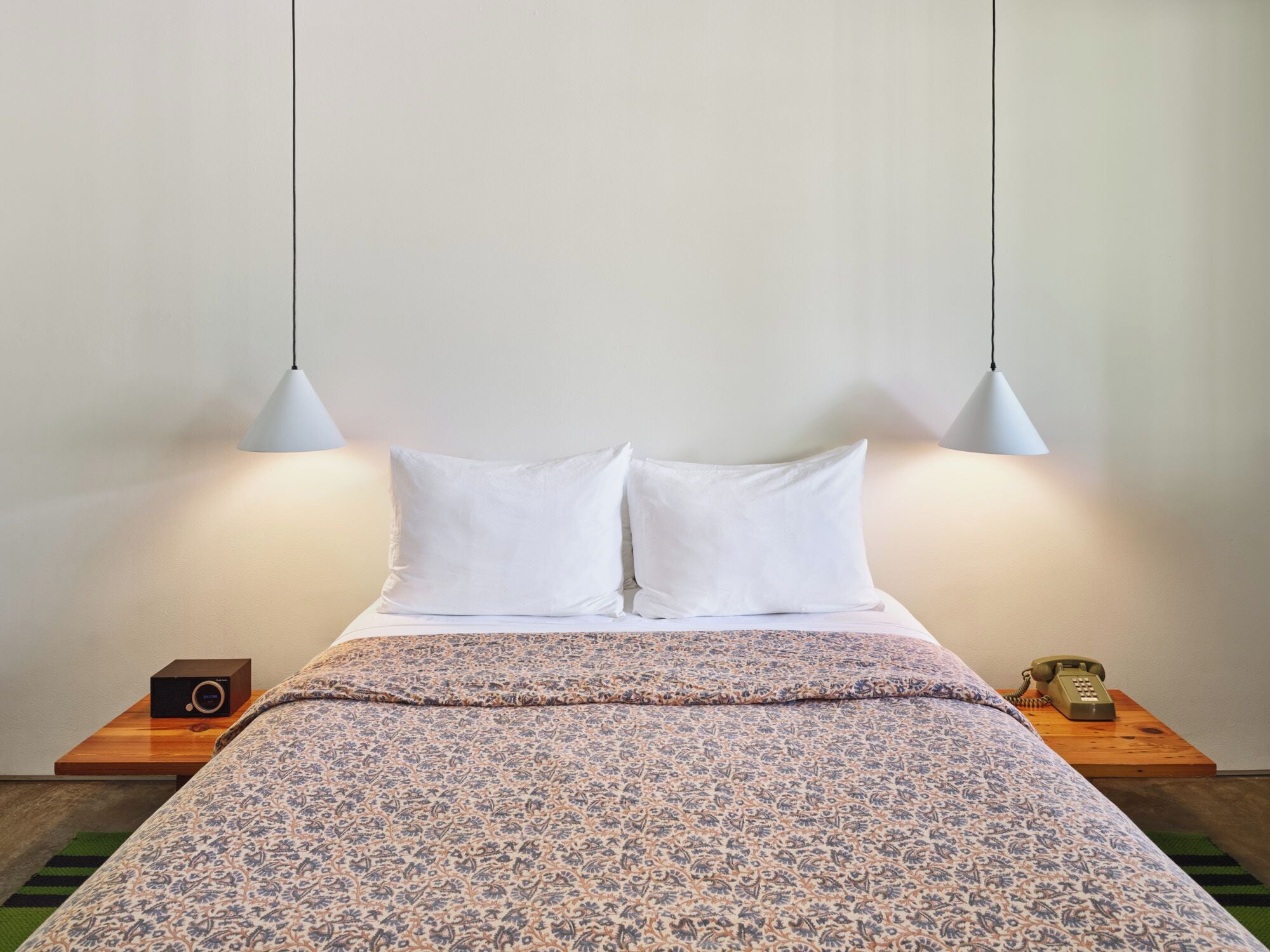 A neatly made bed with a patterned bedspread, two white pillows, and two pendant lights hanging above. There is a wooden nightstand with a radio and a telephone on it.