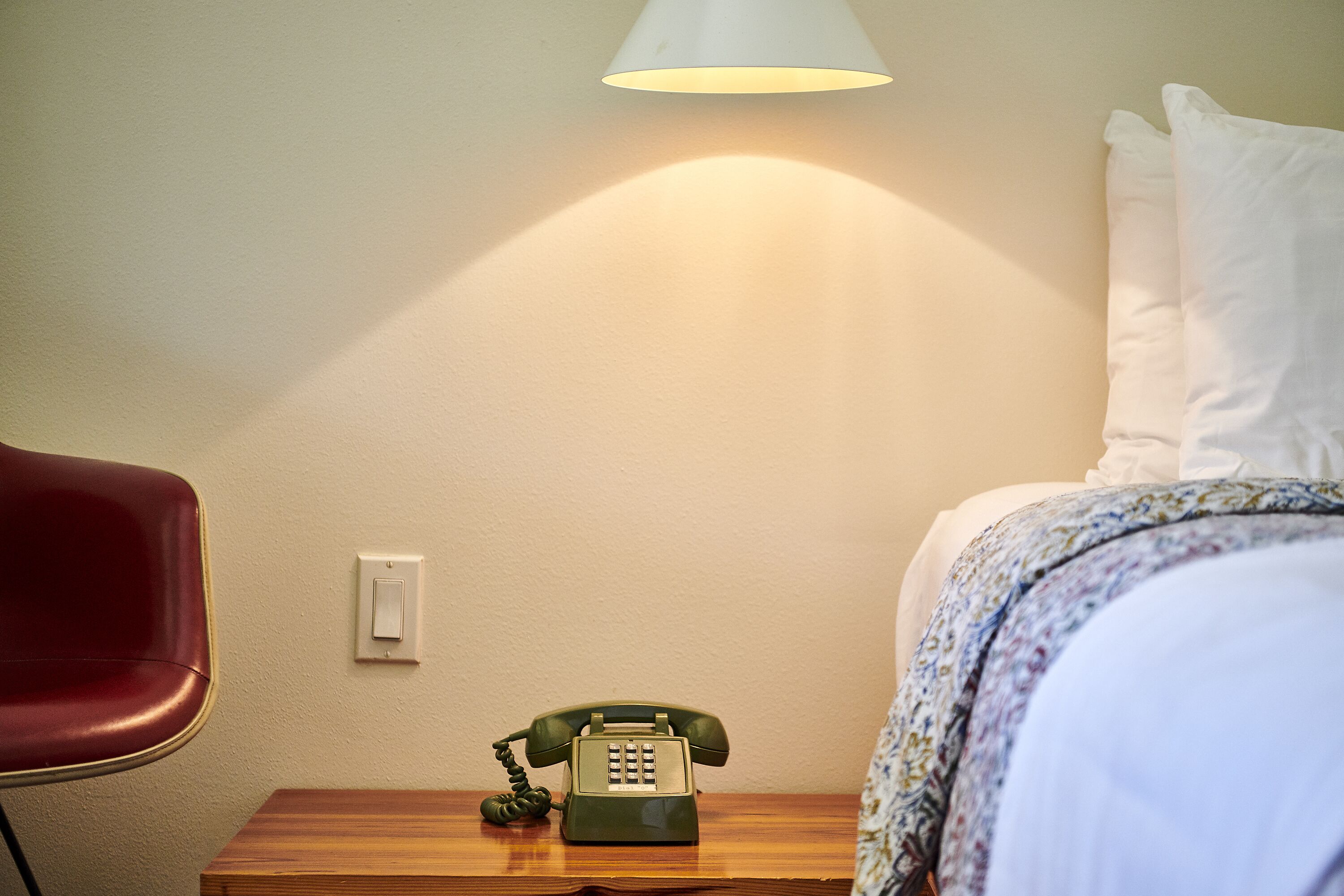 Une chambre d’hôtel avec une table de chevet en bois, un téléphone à cadran vert, une chaise rouge et un lit avec du linge de lit blanc.