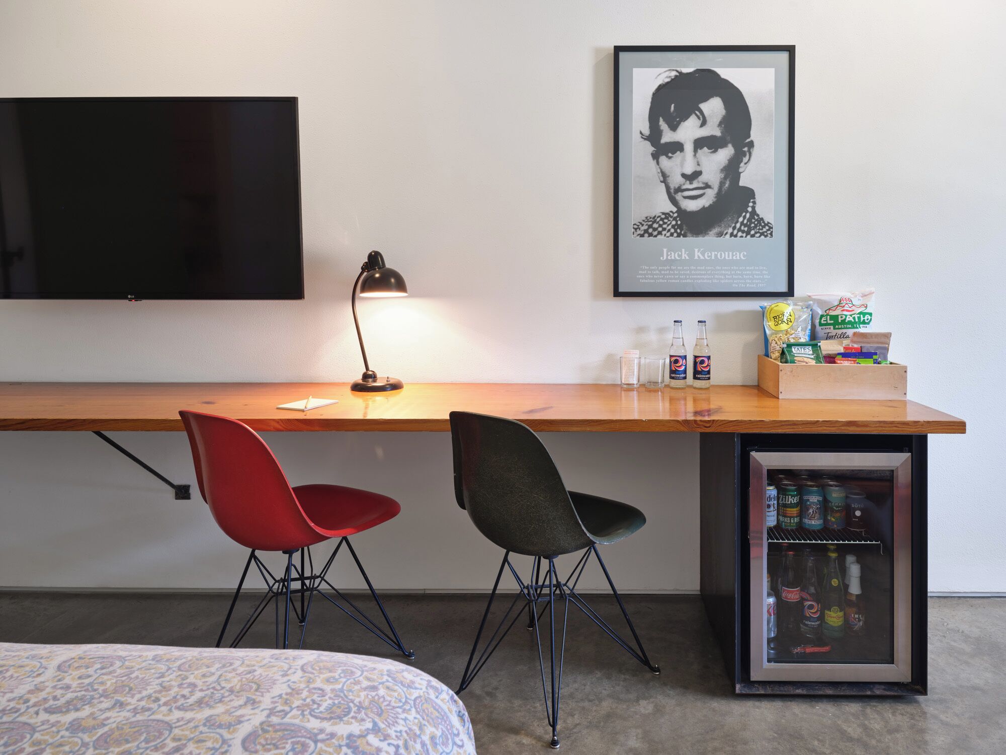 Habitación de hotel con un escritorio de madera, dos sillas, un televisor, un cartel, un refrigerador con bebidas y una cama con cubrecama estampado.