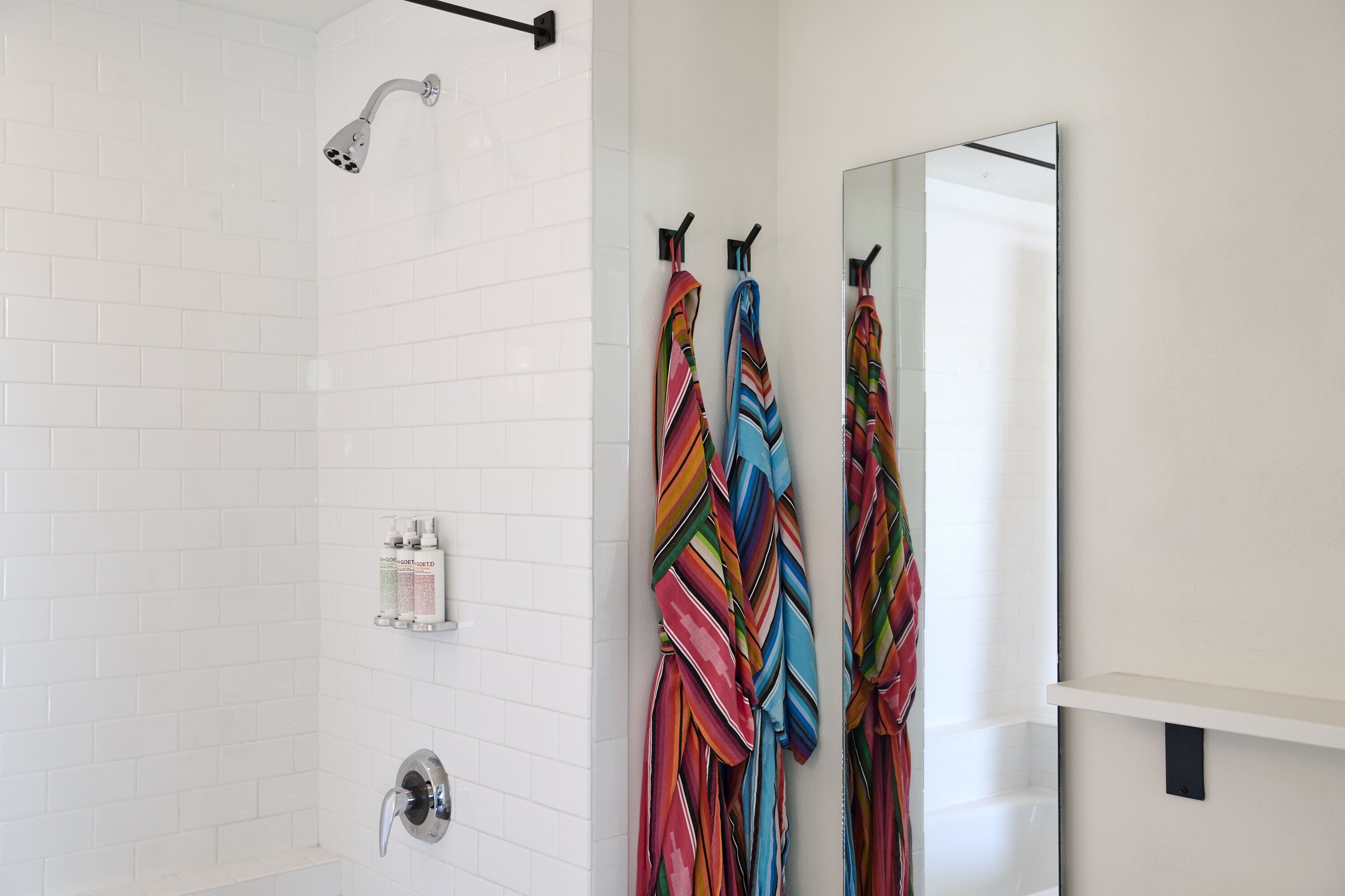 A modern bathroom with white tiled walls, a shower with a handheld showerhead, and a mirror with a towel rack holding colorful towels.