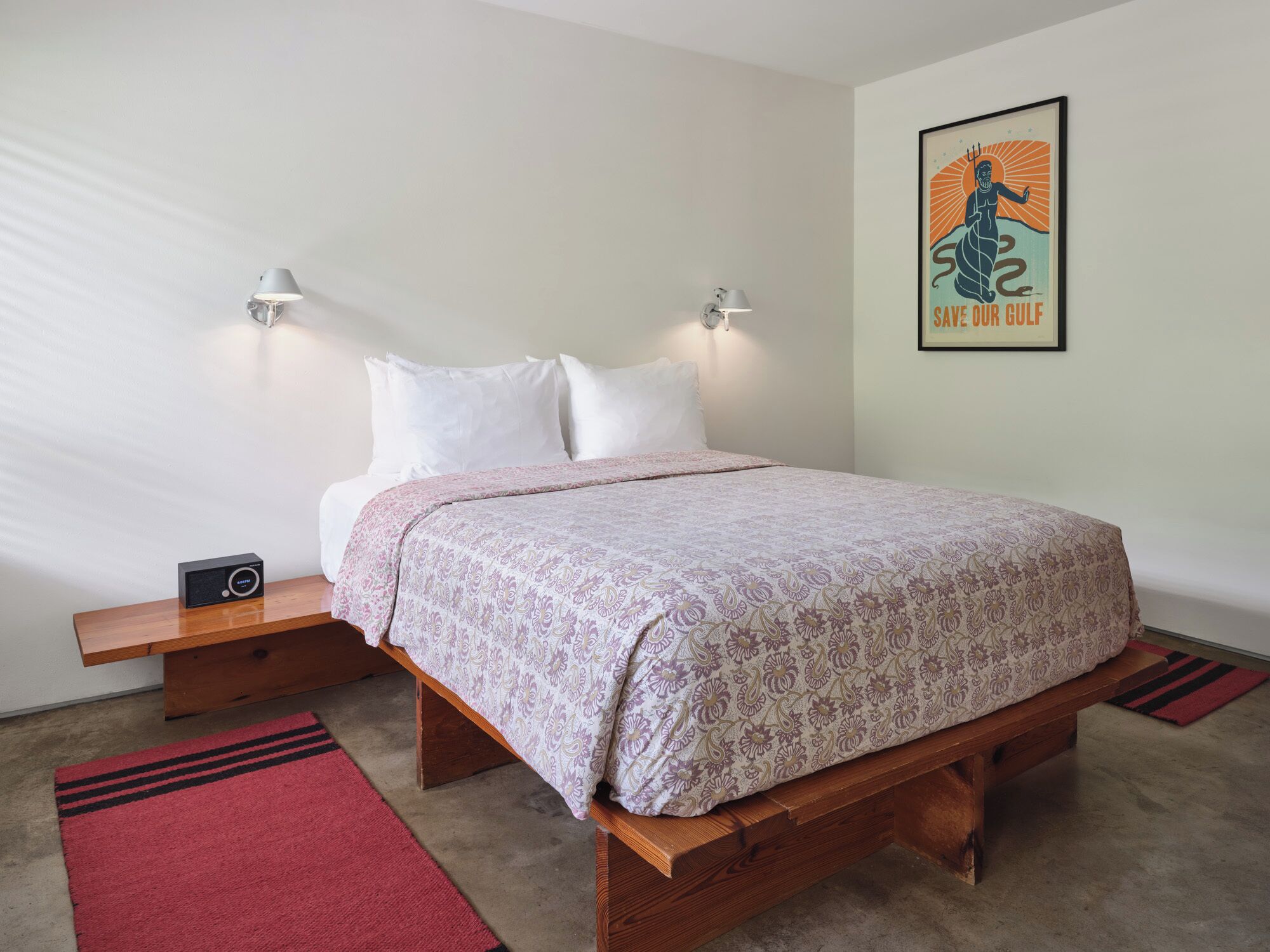 Un lit bien fait, recouvert d’une couvre-lit à motifs, deux tables de chevet avec des lampes, un tapis rouge et une affiche encadrée au mur.