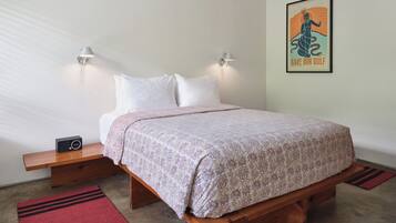 Un lit bien fait, recouvert d’une couvre-lit à motifs, deux tables de chevet avec des lampes, un tapis rouge et une affiche encadrée au mur.
