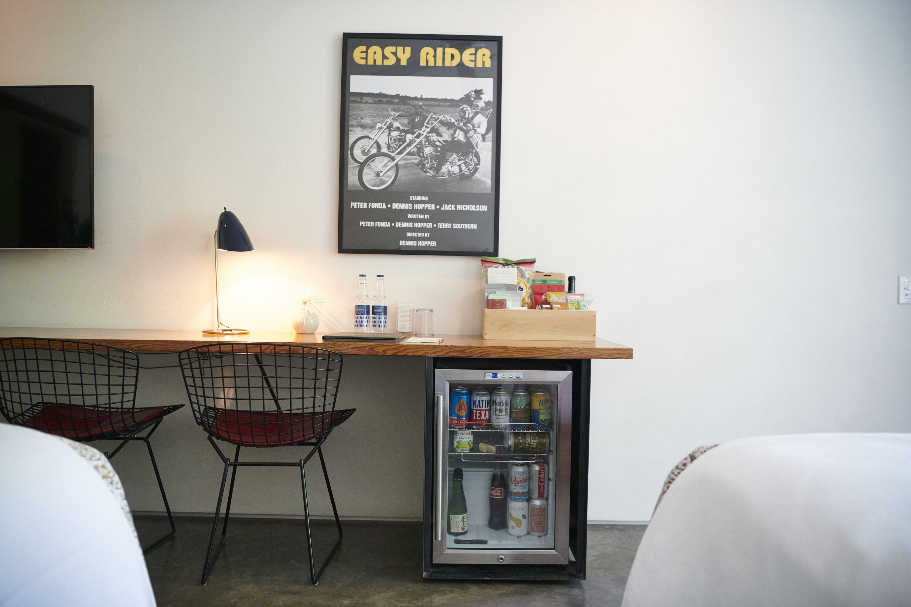 Ett kompakt hotellrum med ett litet skrivbord, två stolar med metallram, ett minikylskåp fyllt med drycker och en affisch av filmen ”Easy Rider” på väggen.