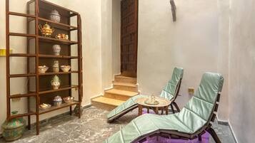 Una habitación con una estantería de madera, un sillón verde acolchado, una pequeña mesa redonda y una escalera que sube a un nivel superior.