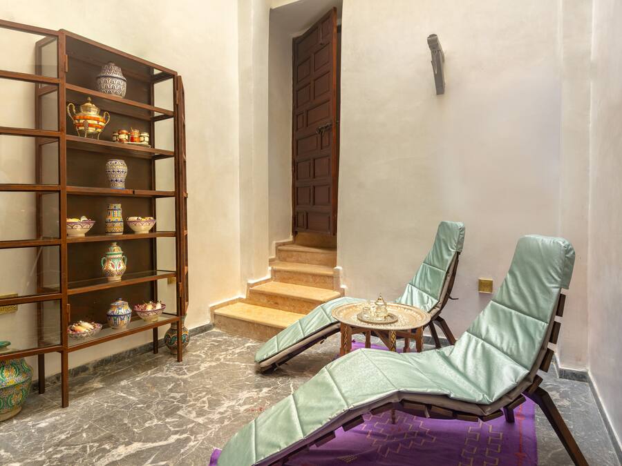 Una habitación con una estantería de madera, un sillón verde acolchado, una pequeña mesa redonda y una escalera que sube a un nivel superior.