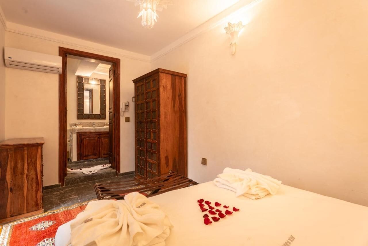 A hotel room with a bed, a wooden wardrobe, a bathroom visible through an open door, and a red carpet.