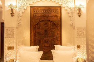 Una habitación con una puerta de madera grande, decorada con elaborados grabados y un diseño de arco tradicional. En primer plano, hay dos almohadas blancas sobre una cama.
