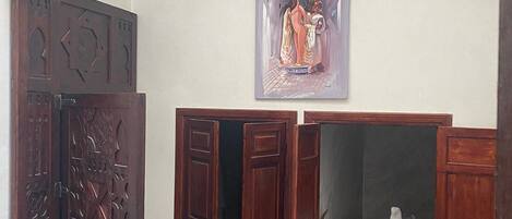 A room with a large wooden door, a painting on the wall, and a small bed with a white cover.