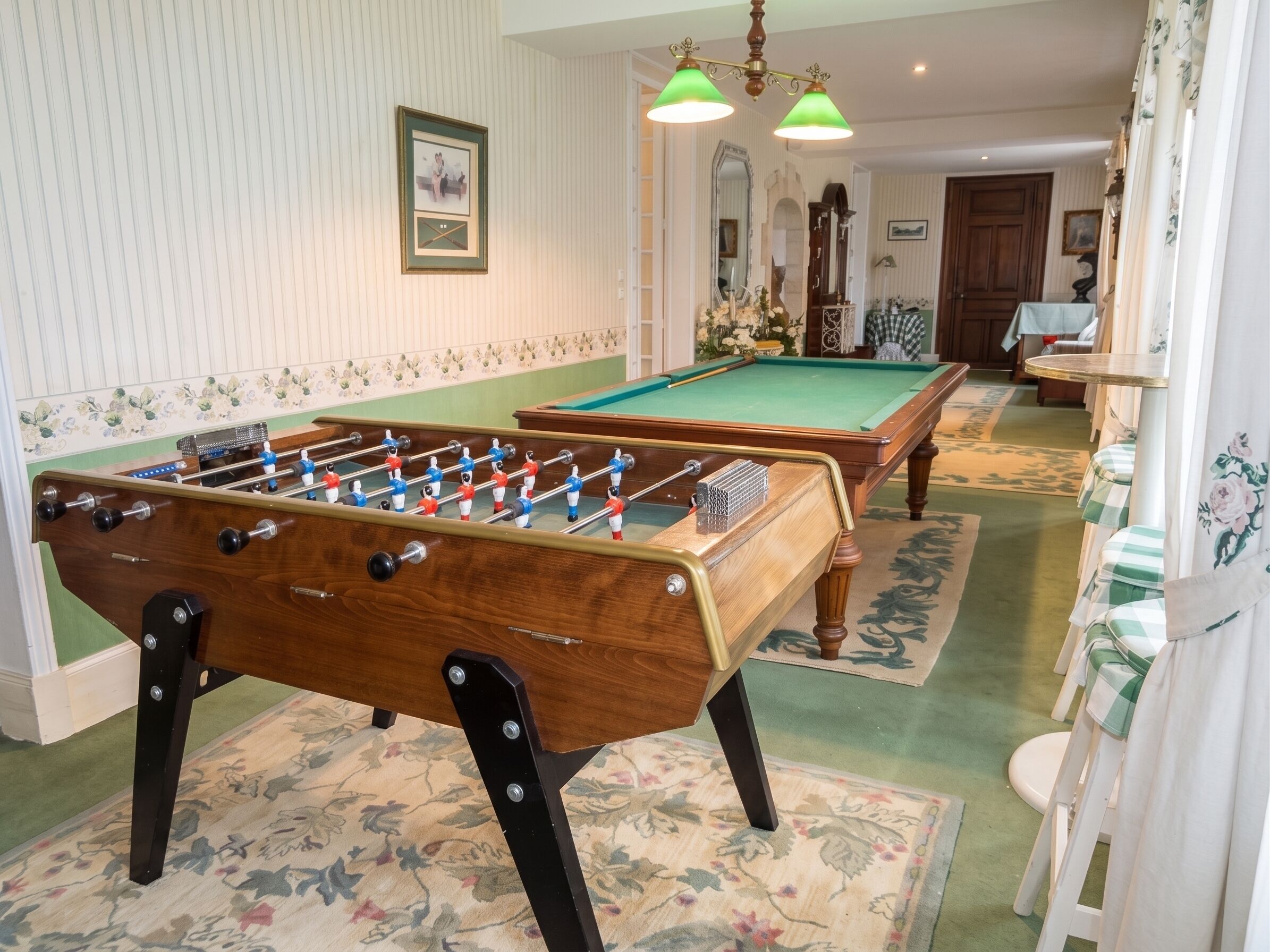 Een pooltafel met een tafelvoetbal in een kamer met gestreept behang, een kroonluchter en een bloemenprint tapijt.