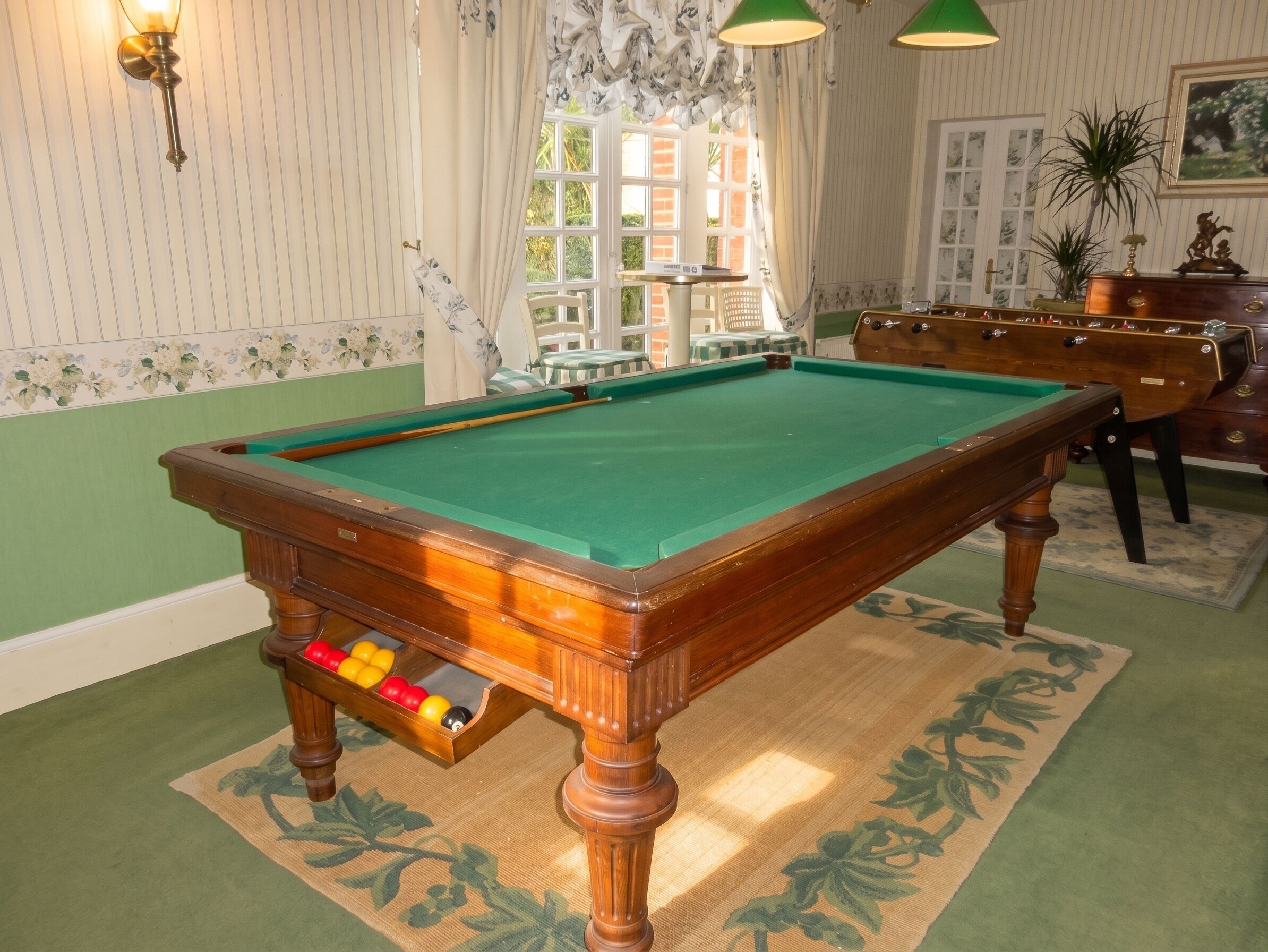 Une table de billard dans une pièce dont les murs sont recouverts d’un papier peint à rayures, avec une moquette verte et une fenêtre ornée de rideaux.