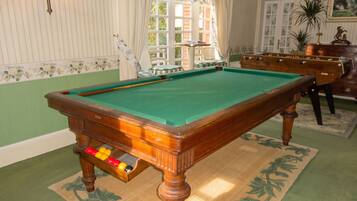 Une table de billard dans une pièce dont les murs sont recouverts d’un papier peint à rayures, avec une moquette verte et une fenêtre ornée de rideaux.