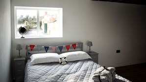 A neatly made bed with a quilted blanket, a bedside lamp, and a small table with a lamp. There is a window with a view of a building outside. - Coastguard Boat House on Belfast Lough (Newtownabbey)
