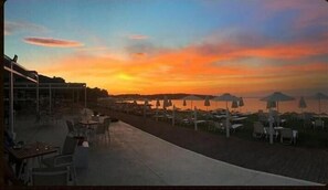 A beachside restaurant with outdoor seating and umbrellas, overlooking a body of water during sunset.