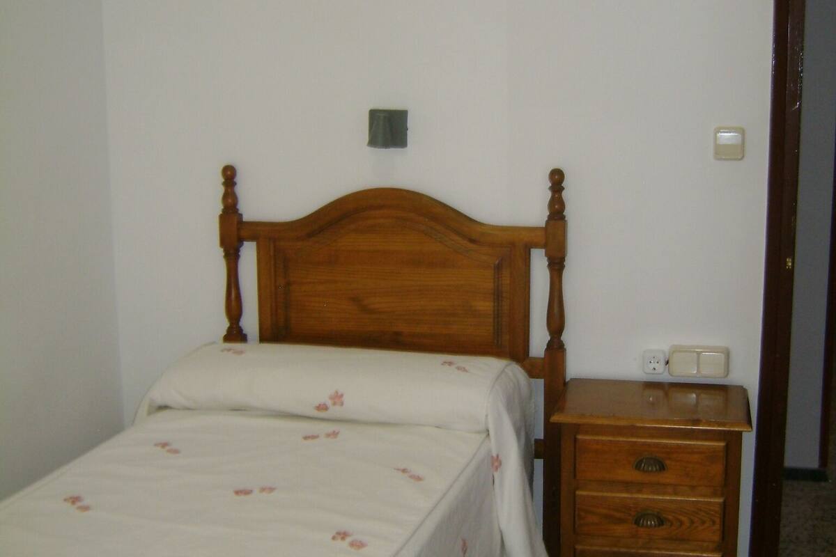 Un lit simple avec une tête de lit en bois, une table de chevet avec une lampe et une petite commode.