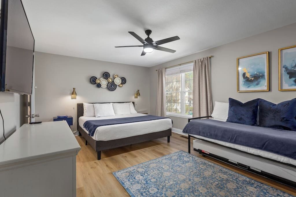 A bedroom with a bed, a sofa, a ceiling fan, a window with curtains, and wall art.