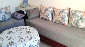 A living room with a grey sofa, floral cushions, a round coffee table with a floral tablecloth, and a small side table with a vase and a handbag. - Duplex feet in water in Taghazout 4563 (Agadir)